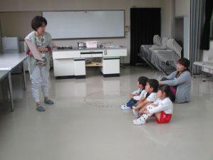 幼稚園4歳児さんが給食センターを見学しています。4歳児さんは3人が地面に座って、職員の話を聞いている写真