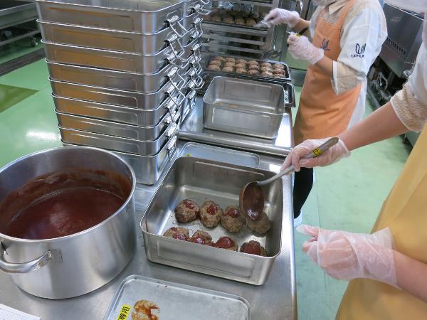 食缶に入れたハンバーグにソースをかける調理員さん